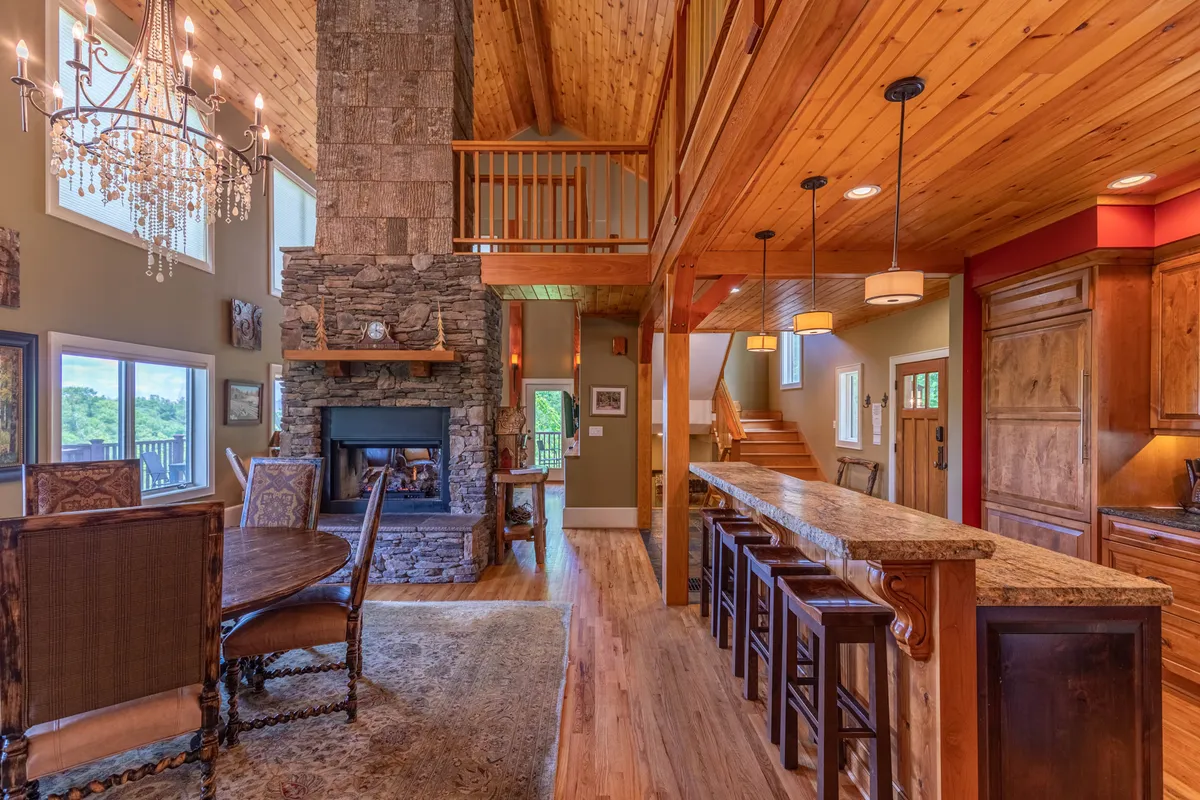 An elegant shot showing a wooden bar area adjacent to a dining table, with a cozy stone fireplace and a warm chandelier overhead. The space is inviting, perfect for sipping cocktails or enjoying a fireside conversation, and highlights the home's hospitality-focused design.