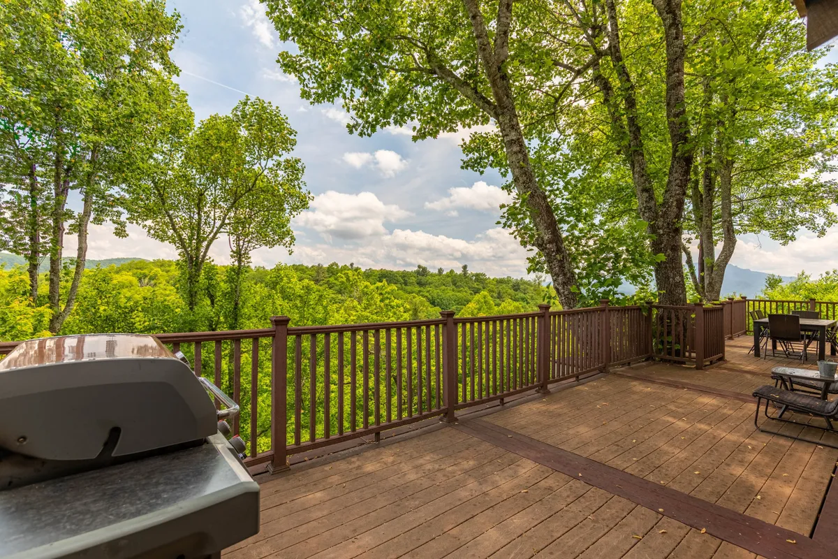A spacious deck nestled among lush greenery, featuring a grill, outdoor dining table, and comfortable seating with panoramic mountain views in the background. The scene is inviting for both daytime and evening relaxation, with ample space for lounging, dining, or soaking in the natural surroundings.