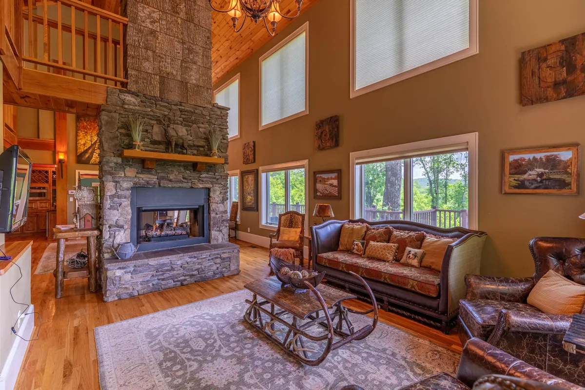 A spacious living area featuring a dramatic stone fireplace that anchors the room, large windows letting in ample light, a wooden staircase, and comfortable seating arranged for conversation or relaxation. The space radiates warmth and invites family and friends to gather around the hearth.