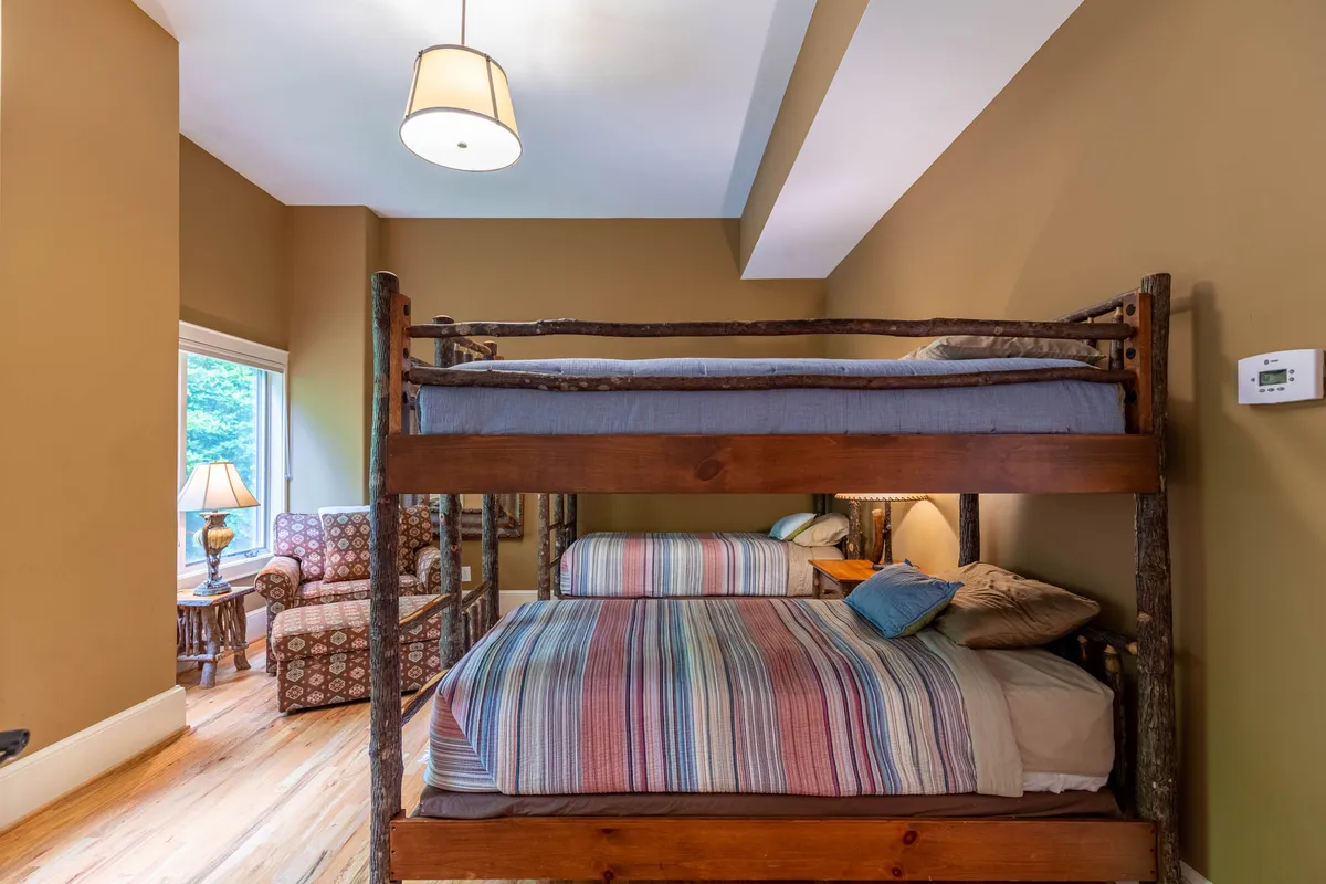 A cozy nook with stacked bunk beds, an armchair, lamp, and natural light streaming through the window. The intimate, inviting space is designed for comfortable nights and quiet moments.