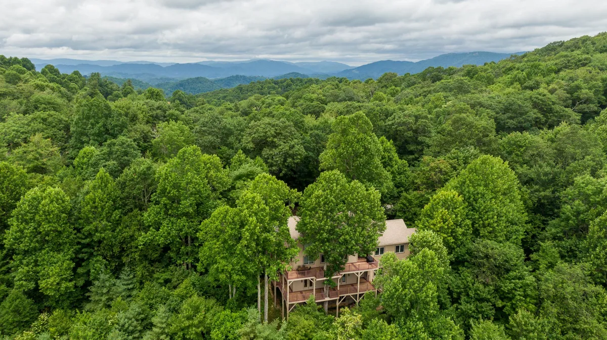 A welcoming mountain house nestled amidst lush trees, with a broad porch and the Blue Ridge Mountains stretching into the background. The home is surrounded by forest, hinting at nearby hiking trails and expansive outdoor adventures.