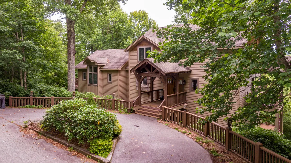 The image showcases the welcoming main entrance of a single-family mountain home, highlighted by a rustic wooden awning. Lush green foliage surrounds the house, and a charming pathway winds invitingly toward the front door, setting a tranquil and picturesque scene.