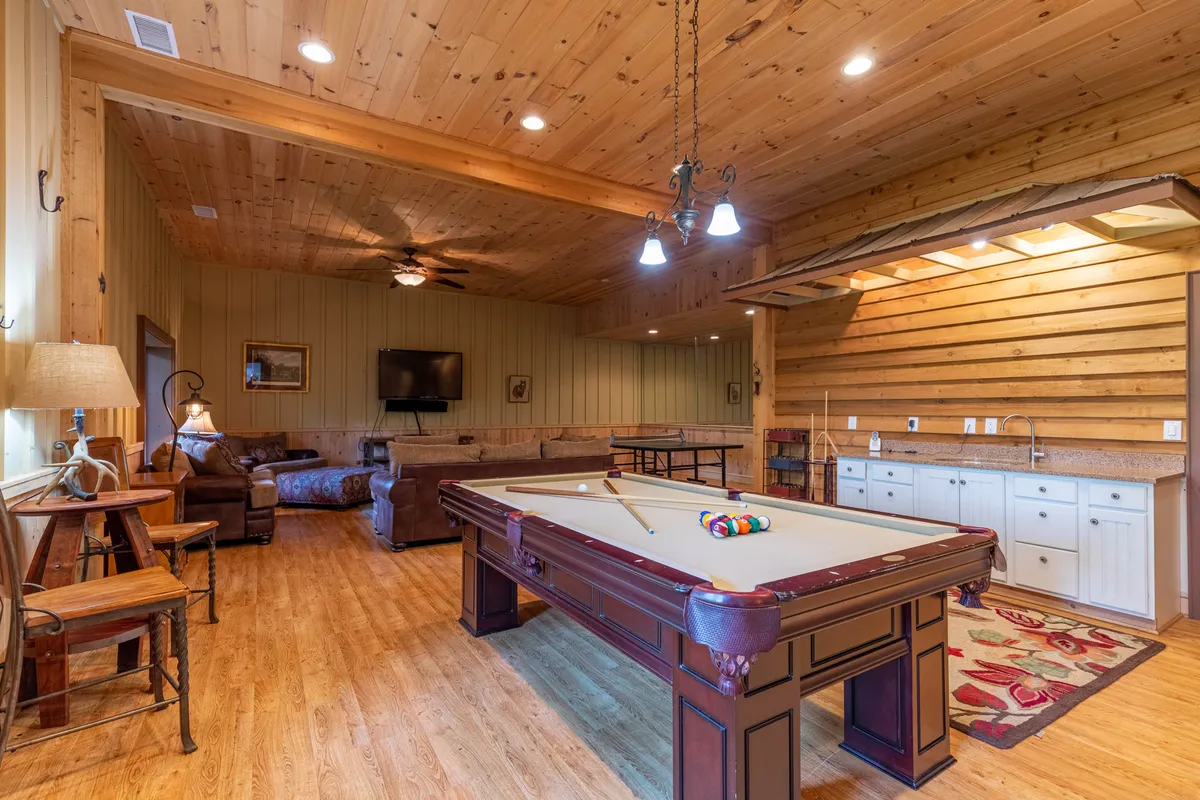 A spacious, relaxed game room featuring a regulation pool table in the foreground, with a comfortable sofa seating area and a convenient kitchenette off to the side. A decorative rug and ambient lighting make the space feel inviting, and the setup is ideal for both playing billiards and socializing.