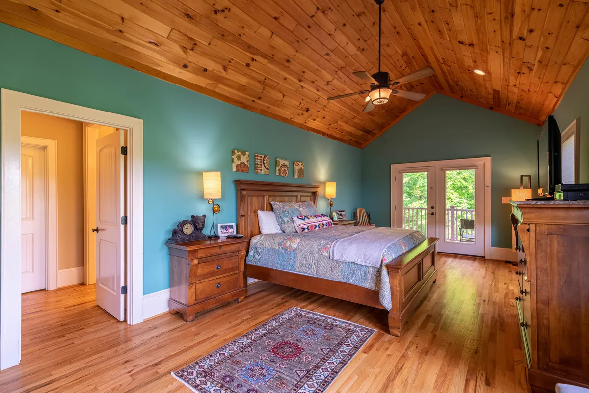 A serene master suite featuring a large, inviting bed, wooden ceiling, and double doors leading to a private balcony. The space is bathed in natural light, creating a peaceful retreat ideal for relaxation and enjoying the view.