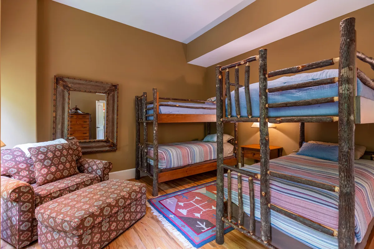 A playful bunk room with multiple bunk beds, striped bedding, and a nature-inspired rug, complemented by a lounge chair. The space is bright and inviting, ideal for kids or cousins to bond and unwind.